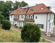 Weißes zweistöckiges Gebäude mit rotem Ziegeldach, mehreren Gauben und Rundbogenfenstern, umgeben von grünen Büschen und Bäumen. Eine kleine gepflasterte Auffahrt und Eingangsstufen mit Blumen flankieren den Eingangsbereich.