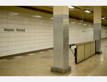 U-Bahn-Station mit einer weiß gefliesten Wand, auf der "Anhalter Bahnhof" steht. Im Vordergrund befinden sich ein rechteckiger Pfeiler aus Beton mit Metallverkleidung und eine Absperrung mit dem Schild "Kein Durchgang".