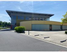 Modernes, eingeschossiges Gebäude mit gelblichen Fassaden und großen Glasflächen, darüber ein auskragendes Flachdach. Im Vordergrund befindet sich ein gepflasterter Parkplatz mit wenigen Begrünungselementen, umgeben von Bäumen und Gehwegen.