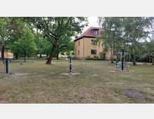 Grüne Parkanlage mit mehreren Fitnessgeräten auf einer Rasenfläche, von Bäumen umgeben. Im Hintergrund stehen gelbe Wohngebäude mit roten Dächern.