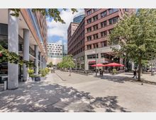 Städtischer Platz mit moderner Architektur, flankiert von Backstein- und Glasgebäuden mit Geschäften und Restaurants. Im Vordergrund befinden sich Bäume, Tische mit roten Sonnenschirmen und Gehwege aus Pflastersteinen.