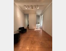 Ein minimalistischer Innenraum mit glatten, warmen, terrakottafarbenen Bodenfliesen, weiß gestrichenen Wänden und einer Deckenbeleuchtung mit mehreren Spotlights. Auf der linken Seite stehen zwei schwarze, niedrige Sessel mit einem kleinen runden Beistelltisch, während im hinteren Bereich des Raumes ein Durchgang mit Tageslicht und Hockern sichtbar ist.