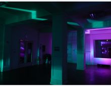 Ein dunkler Innenraum mit weißen Wänden und Säulen, der von farbigem Licht in Lila- und Grüntönen beleuchtet wird. Im Hintergrund sind Türen, ein Fenster und einzelne Möbelstücke erkennbar, die von den Lichtstrahlen angestrahlt werden.