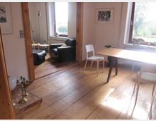 Wohnraum mit Holzboden, Holzbalken und einem Tisch mit weißen Stühlen. Im Hintergrund befindet sich eine Sitzecke mit schwarzem Sessel und Holzfenstern, die Tageslicht hereinlassen, und dekorativen Elementen auf der Fensterbank.