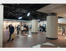 U-Bahn-Station mit Personen, die sich durch einen offenen Bereich mit rötlich-braunem Boden bewegen. Große Säulen mit geriffelter Optik stützen die Decke, die sichtbare Verkabelung und eine abgenutzte Oberfläche aufweist. Hinweise wie ein Schild zur Linie U8 und digitale Anzeigetafeln sind sichtbar.