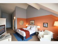 Hotelzimmer mit Doppelbett, oranger Wandakzent, weißen Möbeln und einem cremefarbenen Sessel neben einem kleinen Tisch. An der Wand hängen Bilder, und in einer Ecke steht ein Flachbildfernseher auf einem Sideboard.