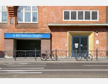 Backsteinfassade mit Zugang zum U-Bahnhof "Rathaus Steglitz", erkennbar an einem blauen Schild. Vor dem Gebäude stehen zwei Fahrräder an einem Gitterzaun, und rechts daneben ist eine Tür mit gelbem Rahmen und Gittertüren sichtbar.