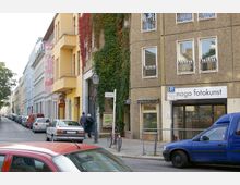 Straßenszene mit mehrstöckigen Wohn- und Geschäftshäusern, darunter ein gelbes Gebäude mit Balkonen und ein mit Efeu bewachsenes braunes Gebäude. Im Erdgeschoss befinden sich Geschäfte, darunter „nago fotokunst“. Auf der Straße parken Autos, und ein Fahrrad steht an einem Laternenmast.