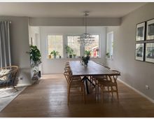 Esszimmer in einem modernen, hellen Haus mit Holzboden und einem großen rechteckigen Holztisch, umgeben von Holzstühlen. Die Wand ist mit vier gerahmten Bildern verziert, und große Fensterfronten lassen viel Tageslicht herein; auf den Fensterbänken und Regalen stehen Pflanzen.