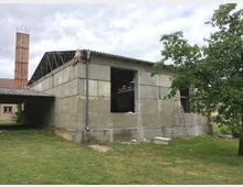 Ein unfertiges oder renovierungsbedürftiges Betongebäude mit offenem Fensterbereich und Wellblechdach. Im Hintergrund sind ein roter Ziegelkamin und ein weiteres Gebäude zu sehen, umgeben von einer Wiese mit vereinzelten Bäumen.