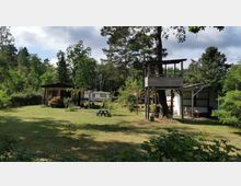Gartenbereich mit Rasenfläche, auf der sich ein Holzspielhaus sowie ein kleiner blauer Picknicktisch befinden. Im Hintergrund stehen ein Holzhäuschen, ein Pavillon und ein Wohnwagen, umgeben von Bäumen und Sträuchern.