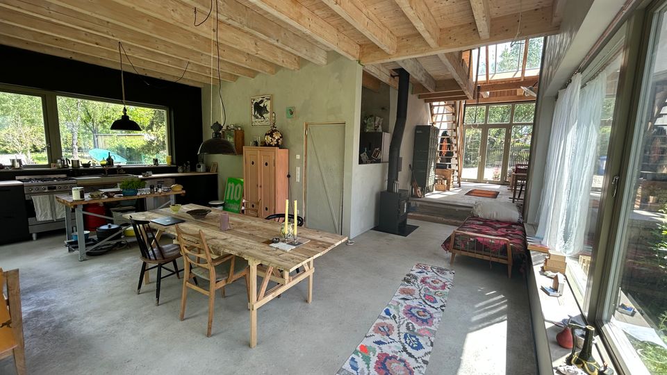 Offener Wohn- und Küchenbereich mit hoher Holzdecke und sichtbaren Balken, großem Esstisch, Küchenzeile und einem Holzofen. Rechts verlaufen bodentiefe Fenster entlang der Wand; im Hintergrund führen Stufen und eine Treppe zu einer verglasten Front mit Blick ins Grün.