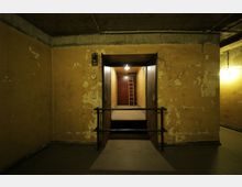 Industrieartiger Raum mit gelben, abgeblätterten Wänden und Sichtbetondecke. Im Zentrum öffnet sich ein beleuchteter Fahrstuhlschacht mit einer Plattform und einer roten Leiter an der Rückwand, umgeben von Metallgeländern.