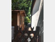 Ein schmaler Weg führt zwischen einem weißen Gebäude rechts und einem hölzernen Gartenschuppen links entlang, umgeben von grünen Bäumen und Sträuchern. Im Vordergrund ist ein Holztor zu sehen, das den Zugang zum Weg abgrenzt.