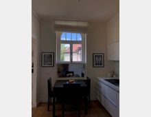 Kleine Küche mit heller Wandfarbe, einem Esstisch mit vier Stühlen und einem großen Fenster mit Rundbogen, das Tageslicht hereinlässt. Auf der rechten Seite befinden sich weiße Küchenmöbel, links an der Wand hängen zwei gerahmte Bilder.