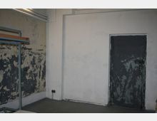 Ein schlichtes, abgenutztes Zimmer mit teilweise abblätternder Wandfarbe; links steht ein einfacher Metallkleiderständer, rechts ist eine dunkle Metalltür mit Farbabplatzungen zu sehen. Die Wände sind in schlichtem Grau und Weiß gehalten, und an der Decke verlaufen Rohre entlang einer Wand.