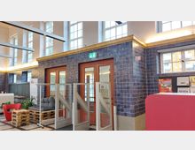 Innenraum eines Gebäudes mit hohen Fenstern, blau gefliesten Wänden und einem Holztürbereich. Im Vordergrund befinden sich Sitzgelegenheiten aus Palettenmöbeln, Pflanzen sowie eine Zugangsschranke.