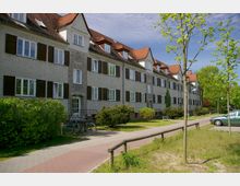 Mehrstöckiges Wohngebäude mit rotgedecktem Satteldach, weißverputzter Fassade und dunklen Fensterläden, umgeben von kleinen Bäumen, Büschen und einem Gehweg im Vordergrund. Links sind ein Fahrrad und rechts ein geparktes Auto zu sehen.