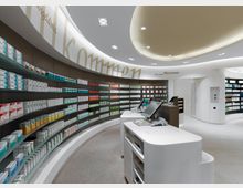 Innenraum einer modernen Apotheke mit gebogenen Regalen, die zahlreiche Arzneimittelpackungen präsentieren. Helle Beleuchtung, einladende Schriftzüge an der Wand und Arbeitsstationen mit Computern im Vordergrund.