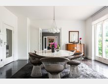 Esszimmer mit modernem, rundem Marmortisch und stilvollen Stühlen aus Drahtgeflecht, einem dunklen Teppichboden und dezenter Beleuchtung durch eine Glas-Pendelleuchte. Im Hintergrund steht ein hölzernes Sideboard vor einer weißen Wand mit Kunstwerken, daneben eine große Fensterfront, die den Raum mit natürlichem Licht füllt.