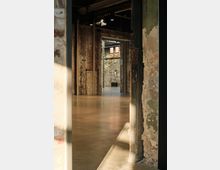 Rauh gestalteter Innenraum eines alten Industriegebäudes mit unverputzten Ziegelwänden, verputzten Bereichen mit abgeplatzter Farbe und einem glatten Betonboden. Tageslicht fällt durch Fenster und eine geöffnete Tür, während der Raum von metallenen Tragstrukturen und Lampen an der Decke durchzogen ist.