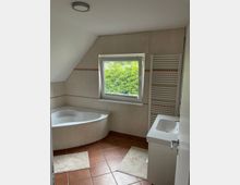 Badezimmer mit Eckbadewanne, einem Waschbecken mit integriertem Unterschrank, einem Heizkörper in Handtuchhalter-Design und einem Fenster, das auf grüne Büsche blickt. Der Raum hat beigefarbene Wände mit roten Akzentfliesen und einen Boden aus rotbraunen Fliesen.