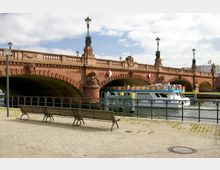 Eine historische Steinbrücke mit mehreren Bögen und dekorativen Skulpturen, darunter Laternenpfosten, überspannt einen Fluss. Im Vordergrund befinden sich eine gepflasterte Uferpromenade mit Sitzbänken und Geländer, während ein weißes Ausflugsschiff unter der Brücke hindurchfährt.