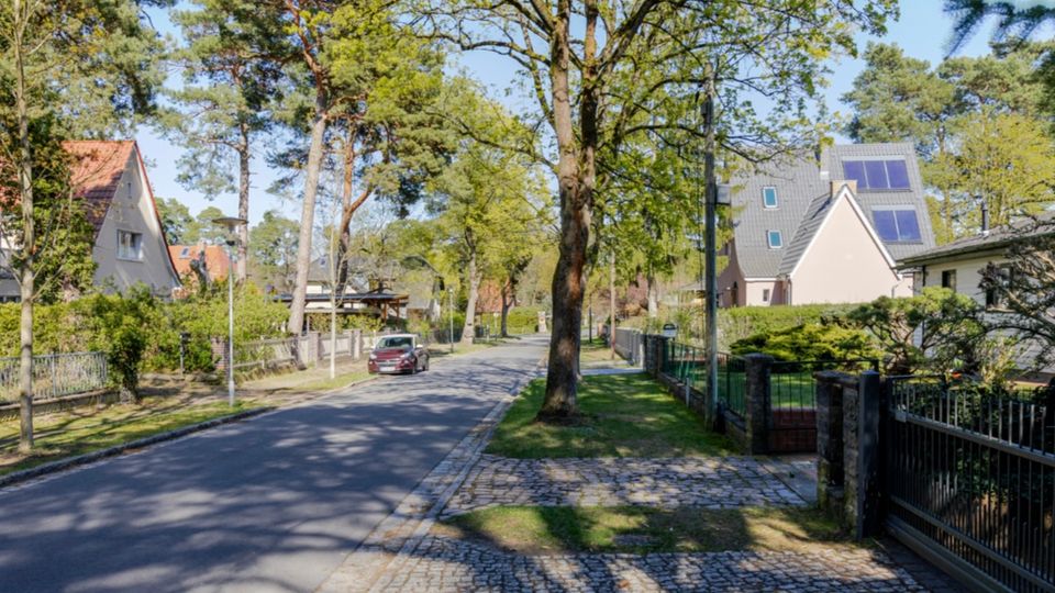 Wohnstraße in einem grünen Wohngebiet mit hohen Bäumen entlang der Fahrbahn, Gehweg und Pflasterstreifen am Rand. Rechts und links stehen Einfamilienhäuser mit Gärten und Zäunen, darunter ein Haus mit Solarmodulen auf dem Dach.