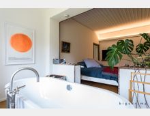 Modernes Badezimmer und Schlafzimmer in einem offenen Raum. Im Vordergrund eine weiße freistehende Badewanne mit Armatur, ein großes Monstera-Blatt auf der rechten Seite, und im Hintergrund ein Bett mit blauer Bettwäsche sowie indirekter Beleuchtung an der Decke.