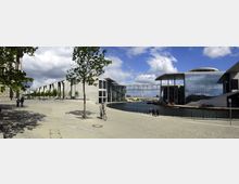 Weitläufiger Platz mit moderner Architektur entlang eines Flusses, geprägt von großen Glasfassaden und einer Fußgängerbrücke, die zwei Gebäude verbindet. Im Vordergrund sind Bäume, Gehwege und eine Person auf einem Fahrrad zu sehen, im Hintergrund der Fluss und weitere Gebäude.