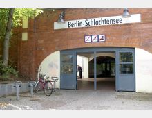 Backsteingebäude mit einer gebogenen Eingangstür, beschriftet mit "Berlin-Schlachtensee". Vor dem Eingang steht ein Fahrrad an einem Fahrradständer, und im Hintergrund ist ein mit grünen Fliesen verkleideter Durchgang sichtbar.