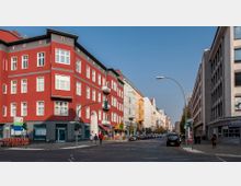 Straßenszene mit einer roten Ecke eines Wohn- und Geschäftshauses, flankiert von mehrfarbigen Altbauten auf der linken Straßenseite und moderneren Gebäuden rechts. Im Vordergrund sind Gehwege, eine Kreuzung, geparkte Autos und einige Passanten zu sehen.