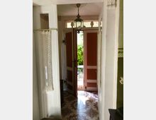 Ein Eingangsbereich mit geöffneten Doppeltüren, die nach draußen führen und einen Blick auf grüne Pflanzen geben. Der Boden ist mit dekorativen Fliesen verlegt, und eine hängende Glaslaterne befindet sich an der Decke.