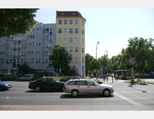 Straßenszene mit mehrstöckigem Wohn- und Geschäftsgebäude im Hintergrund, links grau und rechts gelb gestrichen. Im Vordergrund fahren Autos auf einer mehrspurigen Straße, während links ein U-Bahn-Schild und rechts ein gelber Linienbus sowie Fußgänger und Radfahrer auf einem Bürgersteig sichtbar sind.