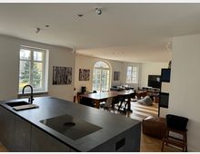 Moderne, helle Wohnküche mit offenem Wohnbereich. Im Vordergrund eine Kücheninsel mit dunkler Arbeitsplatte und integriertem Herd, im Hintergrund ein Essbereich mit großem Holztisch sowie ein Wohnzimmer mit Sofa, Flachbildfernseher und großen Fenstern, die Tageslicht einlassen.