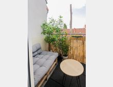 Kleiner Balkon mit einer Sitzecke aus Paletten und grauen Polstern, einem runden Holz-Tisch und mehreren Pflanzen in Töpfen. Der Balkon ist mit einem Sichtschutz aus Bambusstäben versehen, im Hintergrund sind Dächer und ein bewölkter Himmel sichtbar.