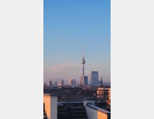 Blick auf den Berliner Fernsehturm und umliegende Hochhäuser in einer urbanen Skyline bei Sonnenuntergang, mit Gebäudedächern und einem Geländer im Vordergrund. Der Himmel ist klar und in blauen und rosafarbenen Tönen gehalten.
