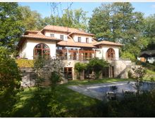 Große Villa im mediterranen Stil mit hellen Wänden, roten Ziegeldächern und zahlreichen großen Fenstern mit Holzrahmen. Die Villa liegt in einer grünen Umgebung mit Bäumen, gepflegtem Rasen und einem leeren Pool im Garten.