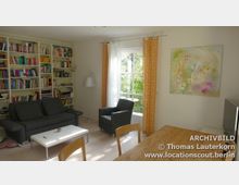 Helles Wohnzimmer mit einer großen Bücherwand, grauem Sofa, Sessel, Couchtisch und Stehlampe. Ein Fenster mit gelben Vorhängen lässt viel Tageslicht herein, daneben ein buntes Gemälde an der Wand.