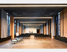 Großer Veranstaltungsraum mit Backsteinwänden und hohen Fenstern, durchzogen von Deckeneinzügen mit moderner Beleuchtung. Der Raum ist bestuhlt, im Hintergrund befindet sich eine kleine Bühne mit zwei Personen.