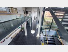 Ein modernes, lichtdurchflutetes Foyer mit hohen weißen Säulen, einer schwarzen Treppe mit Glasgeländern und einer Galerie im Obergeschoss. Große Fenster bieten Sicht auf einen begrünten Außenbereich mit gepflasterten Wegen und Bänken.