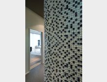 Ein Innenraum mit einer bogenförmigen Wand aus gläsernen, transparenten und dunklen Mosaikelementen im Vordergrund. Im Hintergrund ist ein moderner Raum mit Holzboden, Teppich und schlichten weißen Möbeln sichtbar.