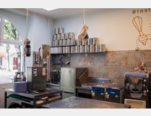 Moderne Gewerbeküche mit Edelstahl-Arbeitsflächen, einer großen Anzahl an Metallbehältern auf einem Regal, industriellen Küchengeräten und einer Fliesenwand aus gemustertem Stein. Ein großes Fenster mit Rundbogen bietet Blick nach draußen.