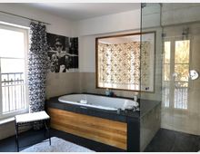 Modernes Badezimmer mit einer freistehenden Badewanne, die in eine Holzverkleidung integriert ist, einer gläsernen Duschkabine, großen Fenstern mit Vorhängen und einem dekorativen Schwarz-Weiß-Poster an der Wand. Der Raum ist hell und schlicht gestaltet, mit Fliesenboden und beigefarbenen Wandfliesen.