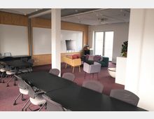 Ein moderner Seminarraum mit violettem Teppichboden, schwarzen Tischen und grauen Bürostühlen. Im Hintergrund befinden sich eine Sitzgruppe mit bunten Sesseln und Sofa, eine große Fensterfront, Whiteboards an der Wand sowie ein Flipchart.