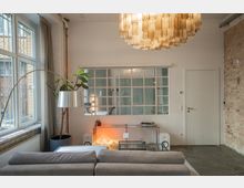 Wohnzimmer mit modernem und industriellem Design, featuring eine große Fensterfront mit Vorhängen links, eine Wand mit freigelegten Ziegeln rechts und eine weiße Wand mit einer Gitterfensterstruktur in der Mitte. Im Vordergrund stehen ein graues Sofa, eine Stehlampe und ein niedriger Glastisch; im Hintergrund befinden sich ein Regal mit Deko-Elementen, ein Plattenspieler und eine auffällige, goldene Hängeleuchte.