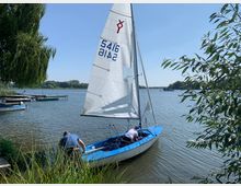 Segelboot auf einem See, mit zwei Personen, die das Boot vorbereiten. Im Hintergrund befinden sich ein Steg mit weiteren Booten, Bäume am Ufer und ein klarer Himmel.