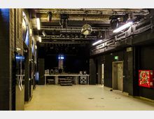 Innenraum eines kleinen Veranstaltungssaals mit schwarzer Bühne im Hintergrund, auf der Musikinstrumente und technische Geräte stehen. Die Wände sind dunkel gestrichen, die Decke trägt Beleuchtungs- und Bühnentechnik, und ein Spiegelball hängt zentral an der Decke.