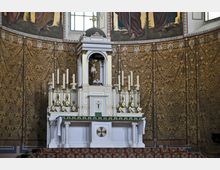 Weißer Kirchenaltar mit goldenen Verzierungen und mehreren Kerzenhaltern, umgeben von einer kunstvoll gemusterten, goldfarbenen Tapete und christlichen Wandgemälden. Oberhalb des Altars befindet sich ein kleines Kruzifix in einer verzierten Nische.