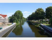 Ein ruhiger Kanal mit Bäumen gesäumten Ufern; auf der linken Seite ein renovierungsbedürftiges Gebäude mit Gerüsten, auf der rechten Seite eine Grünfläche mit Wegen und Sitzbereichen. Ein Boot ist in der Ferne auf dem Wasser sichtbar.